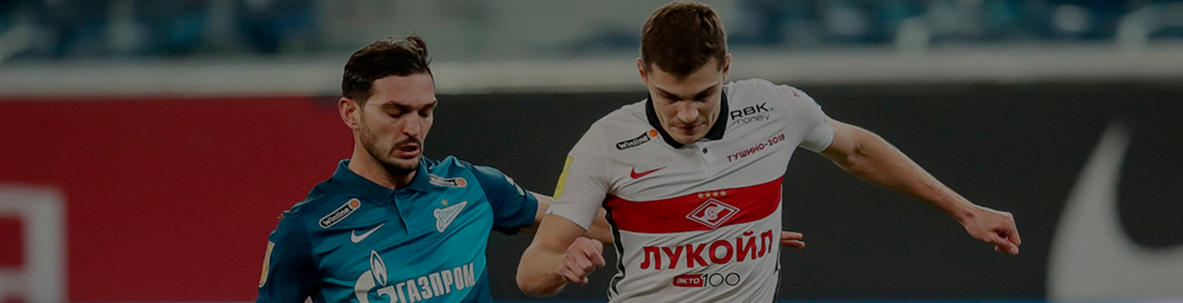 На изображении два футболиста в игровой ситуации. Один в синей форме, другой в белой с красной полосой. Они борются за мяч на фоне футбольного поля и размытых трибун