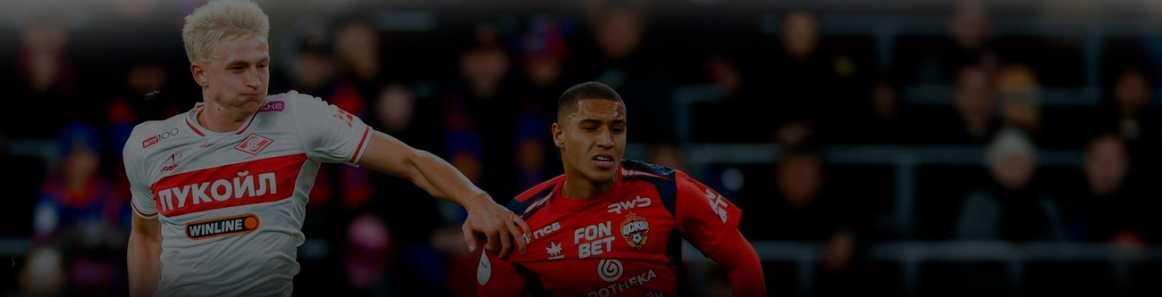 На изображении два футболиста в момент активной игры. Игрок в белой форме с эмблемой «Лукойл» взаимодействует с соперником в красно-синей форме. На заднем плане трибуны стадиона